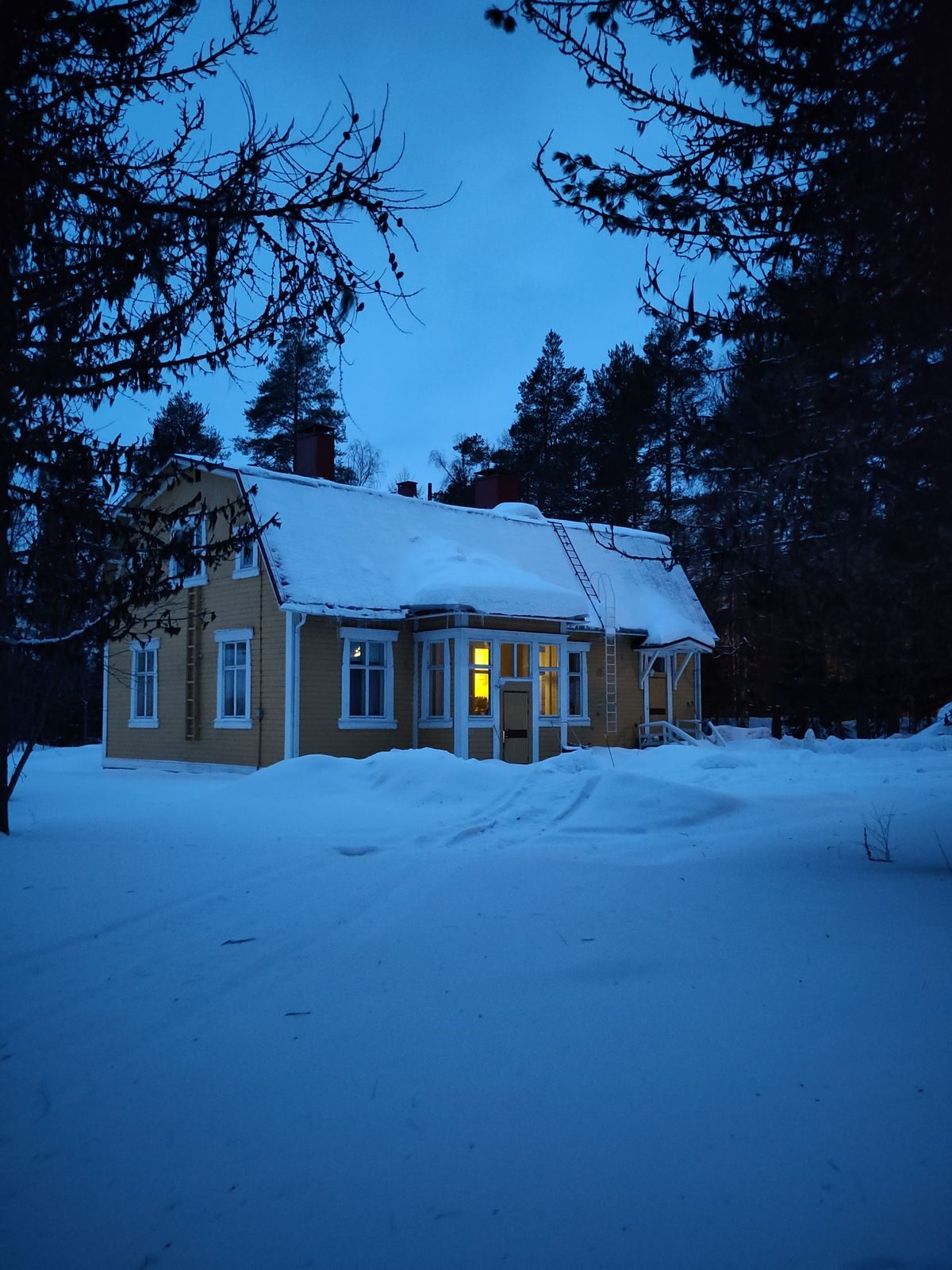 Vaalea omakotitalo lumisessa metsässä hämärän aikaan, yhdessä ikkunassa valo.