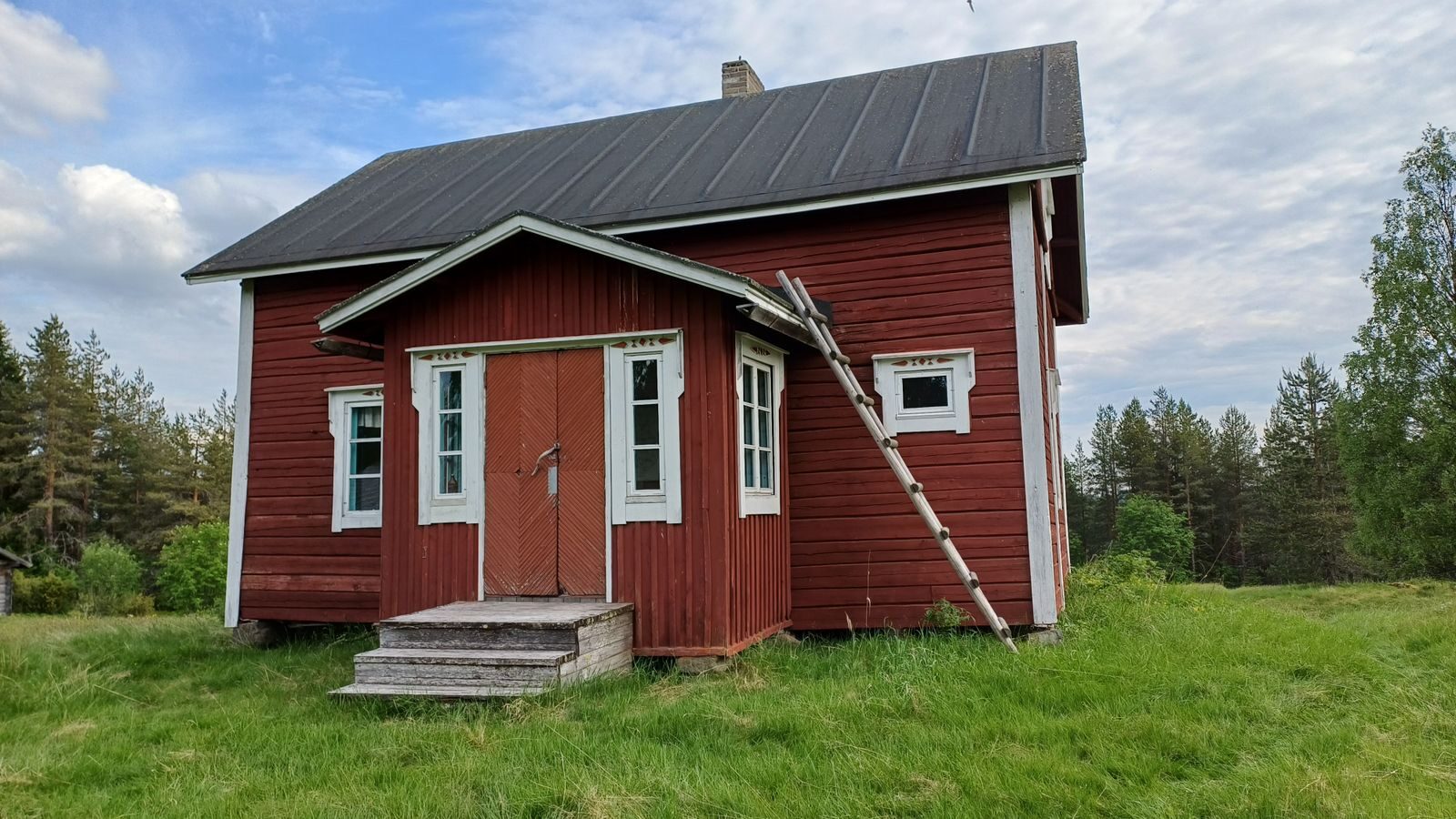 Punainen puutalo nurmipihalla metsän reunassa.