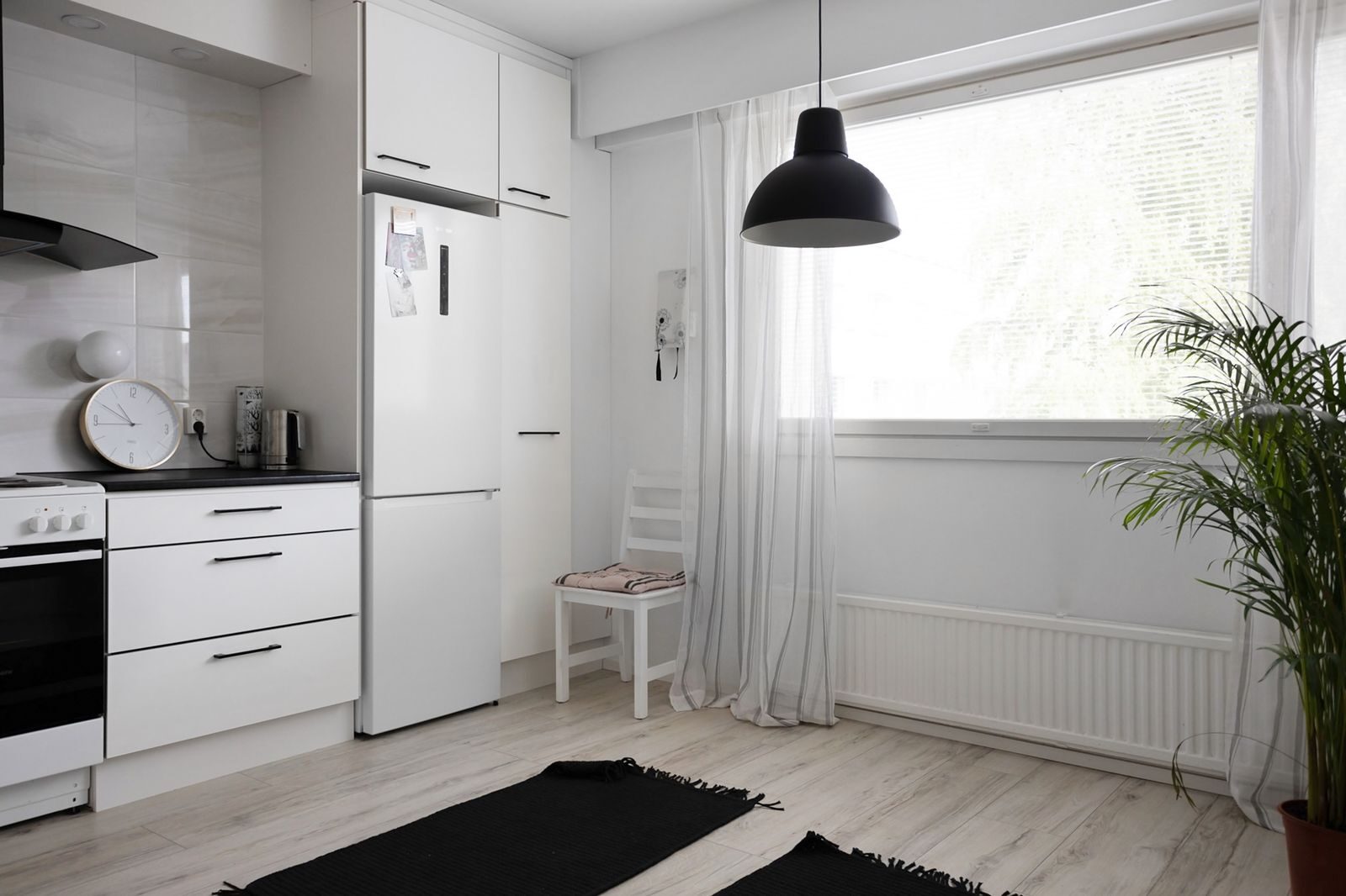 Vaalea keittiö, jossa on sähköliesi, jääkaappi ja ikkunan edessä riippuvalaisin.