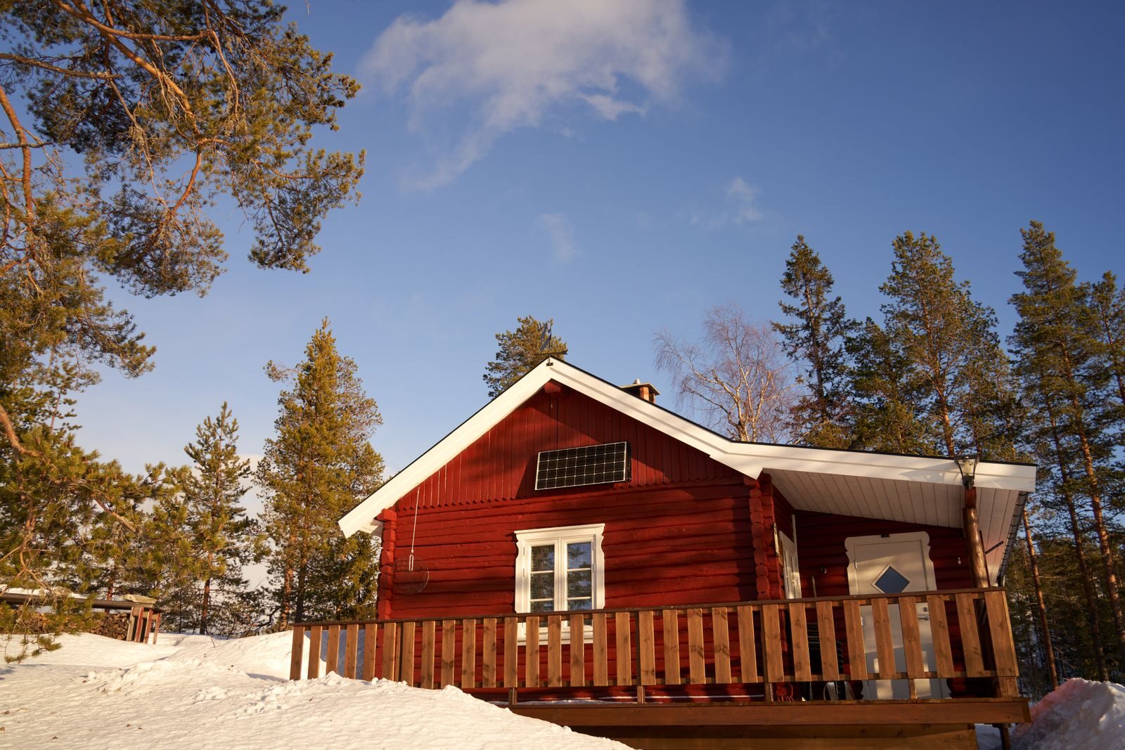 Punainen mökki lumisessa metsässä ja pieni aurinkopaneeli päätyseinällä.