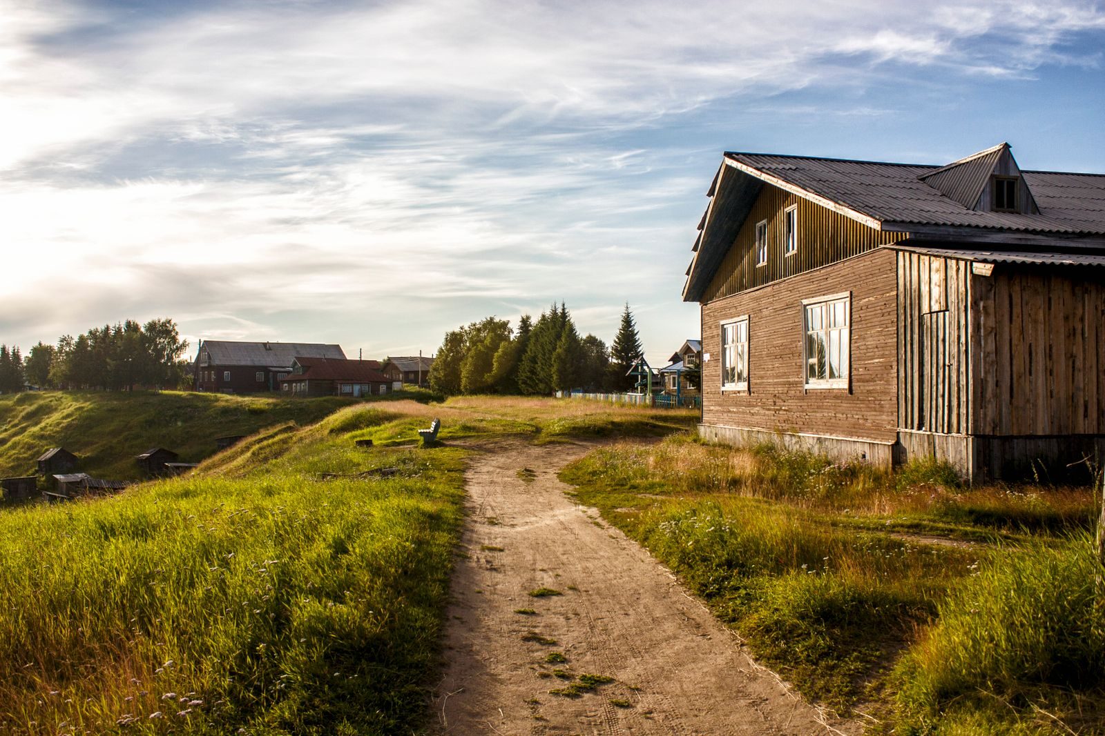 Puutalo ja hiekkapolku maaseutumaisemassa.